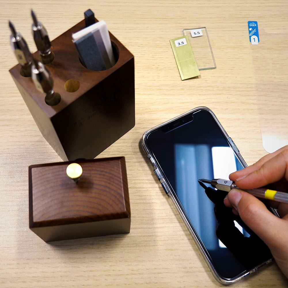 birds eye view of desk with scratch pen tool set in dark wooden box. A hand is holding a scratching scribe and is testing scratch resistance of an Eiger screen protector on a mobile phone
